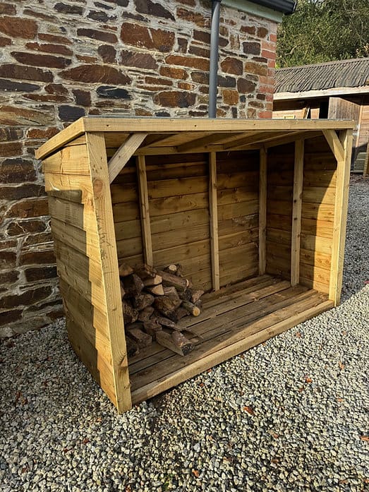 A slatted timber log store stacked with firewood beside a house wall